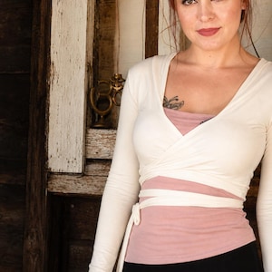 May include: A cream-colored wrap top with long sleeves, layered over a pink bandeau and a black skirt. The top has a wrap design with a tie at the waist. The background is a wooden door.