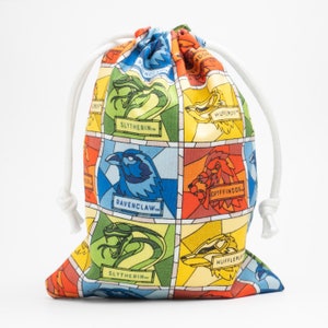May include: A colorful drawstring bag with a stained glass design featuring the Hogwarts house crests: Gryffindor, Hufflepuff, Slytherin, and Ravenclaw. The bag is made of fabric and has white drawstrings.