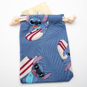 May include: A blue drawstring bag with a pattern of Stitch from Lilo & Stitch surfing on a surfboard. The bag has a light blue background with a palm leaf pattern. The bag is filled with a gift card that says "$100".