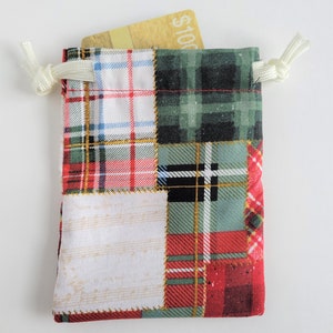 May include: A small, patchwork fabric drawstring bag with a gold gift card peeking out. The bag features a mix of red, green, and white plaid patterns. The drawstring is white.