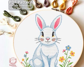 May include: A cross-stitch pattern featuring a white bunny with pink inner ears, sitting on green grass with colorful flowers. The design is in a wooden embroidery hoop, with various colored embroidery floss skeins around it. Text reads "PDF Pattern Instant Download 300 x 300 Stitches."
