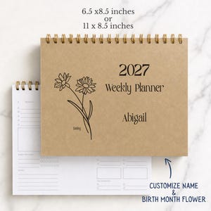 May include: A 2027 weekly planner with a tan cover, gold spiral binding, and a black line drawing of a daisy. The text reads 2027 Weekly Planner and Abigail. Available in 6.5 x 8.5 inches or 11 x 8.5 inches. Includes a white interior page layout.