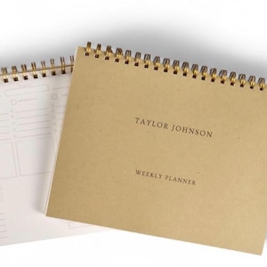 May include: Two spiral-bound weekly planners. One planner is open, displaying a white interior with a grid layout. The other planner has a tan cover with the text "TAYLOR JOHNSON WEEKLY PLANNER" in a simple font. The spiral binding is gold.