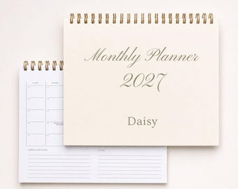 May include: A cream-colored spiral-bound monthly planner for 2027 with the name Daisy printed on the cover. The planner is open to show a grid layout for monthly dates and a section for important dates and notes with black text on white paper.