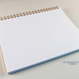 May include: A white dot grid notebook with a gold spiral binding. The notebook is open, displaying the blank pages with a subtle dot grid pattern. The text "DOT GRID SHEETS ON BACK FOR NOTES" is visible.