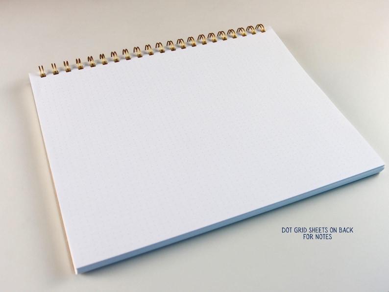 May include: A white dot grid notebook with a gold spiral binding. The notebook is open, displaying the blank pages with a subtle dot grid pattern. The text "DOT GRID SHEETS ON BACK FOR NOTES" is visible.