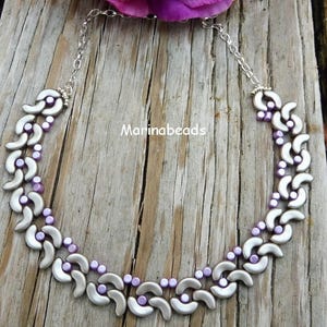 Könnte beinhalten: Eine silberne und lavendelfarbene Perlenkette mit einem Halbmondmuster. Die Kette liegt auf einer Holzoberfläche.
