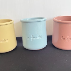 May include: Three ceramic pots in yellow, light blue, and pink, each featuring a smooth, rounded silhouette and a flared rim. The text "la fermière" is embossed in a cursive font on the front of each pot, which has a glossy finish.