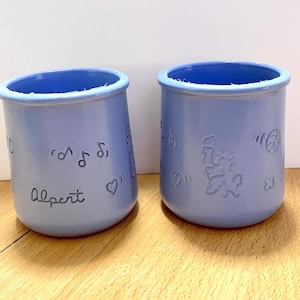 May include: Two light blue ceramic containers with a slightly textured surface. One has the name "Albert" and musical notes etched into the side. The other has a design of a bear, musical notes, and a heart. The rims are a darker blue.