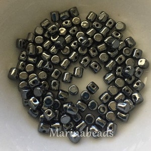 May include: A close-up of a bowl filled with approximately 100 small, dark gray, metallic beads. The beads are round with a flat top and a hole in the center. The beads are arranged in a random pattern.
