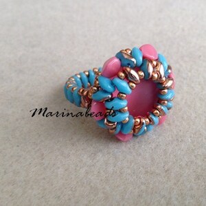 Puede incluir: Un anillo hecho con cuentas azules y rosas, con una piedra rosa en el centro. El anillo est&aacute; hecho con una t&eacute;cnica de tejido de cuentas.