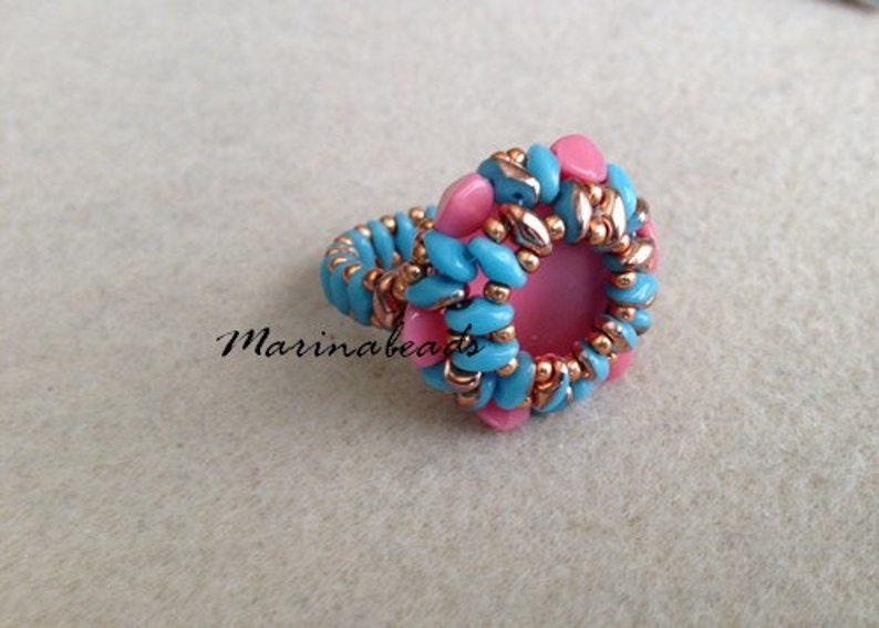 Puede incluir: Un anillo hecho con cuentas azules y rosas, con una piedra rosa en el centro. El anillo est&aacute; hecho con una t&eacute;cnica de tejido de cuentas.