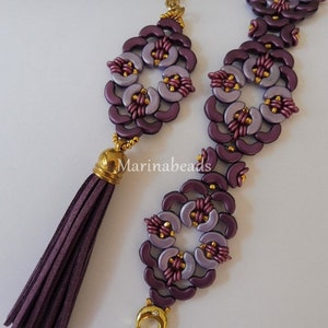 Op de afbeelding: Een paars-gouden kralenset met een ketting en armband met een kwast. De ketting heeft een gouden ketting en een paarse kwast. De armband heeft een gouden sluiting.