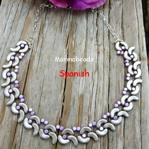 May include: A silver and purple beaded necklace with a curved design. The necklace is on a wooden surface. The text "Marinabeads Spanish" is visible in the image.