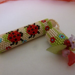 May include: A beaded cylindrical container and lid, decorated with red ladybirds, blue and red flowers, and green accents. The lid is adorned with small flowers and leaves. The container is beige and green.