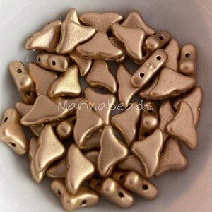 May include: A close-up of a pile of gold-colored glass beads in the shape of a triangle with rounded edges. The beads have two holes each.