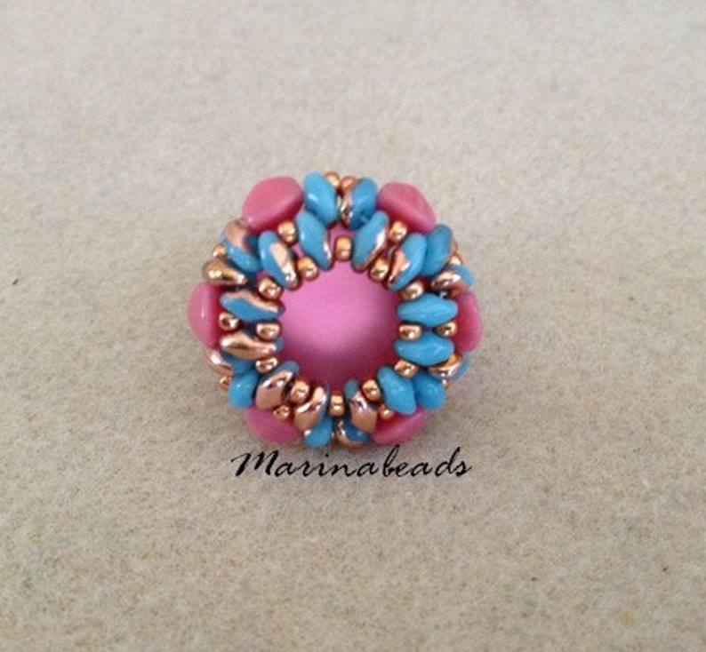 Puede incluir: Un anillo de cuentas rosa y azul con un dise&ntilde;o de alambre de cobre. El anillo est&aacute; hecho de peque&ntilde;as cuentas redondas y tiene una forma circular.
