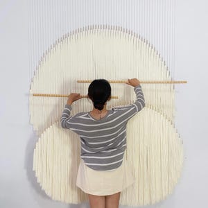 May include: A large wall hanging made of off-white cotton cord arranged in two overlapping circular patterns. The piece is suspended from a horizontal wooden dowel at the top, with two additional wooden dowels positioned horizontally across the centre.
