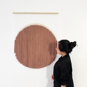 May include: A large, round wall hanging with a textured, rust-colored center. The piece is suspended from a natural wood dowel with white cords and a semi-circle of tan accents. The artwork is displayed against a white wall.
