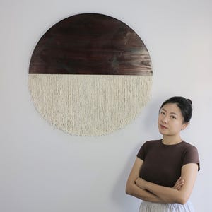 May include: A large, round wall hanging with a dark brown wooden top half and a cream-colored macrame fringe bottom. The wall art is mounted on a white wall, creating a striking contrast. The piece adds a touch of bohemian style to any space.