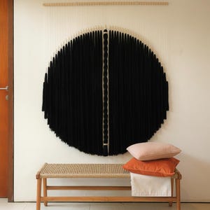 May include: A large, circular black wall hanging with a vertical white stripe down the center. A wooden bench with a woven seat holds three pillows in shades of pink, orange, and white. The wall hanging is suspended from a wooden rod.