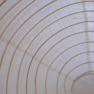 May include: A white fabric lampshade with a circular structure featuring concentric wooden ribs. The material is semi-translucent, allowing light to diffuse through the weave. The word TALANA is printed in small, dark letters on the fabric surface.