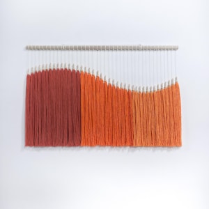Peut inclure: Une tenture murale avec un dégradé de fils de laine orange et rouge. Les fils sont suspendus à une baguette en bois clair, créant un motif texturé en forme de vague. Le fond est un mur blanc uni.