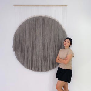 Op de afbeelding: Een grote, grijze, cirkelvormige wandhanger gemaakt van verticale strengen, opgehangen aan een houten staaf. Het kunstwerk is op een witte muur gemonteerd. Het stuk heeft een textuur en creëert een focuspunt in de kamer.