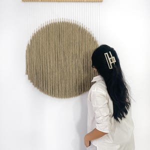 Könnte beinhalten: Ein beigefarbener Wandbehang mit kreisförmigem Design, aufgehängt an einer Naturholzstange. Das Kunstwerk zeigt ein Sonnenmuster, das mit vertikalen Garnsträngen geschaffen wurde und eine strukturierte, minimalistische Ästhetik erzeugt.