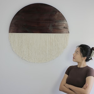 Puede incluir: Un adorno de pared circular, con una sección superior de madera marrón oscuro y una sección inferior de flecos color crema. La pieza está montada en una pared blanca. Este artículo decorativo añade un toque bohemio y mide unos 60 cm de diámetro.