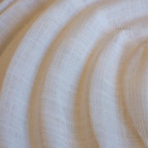 May include: A sheer, off-white fabric featuring a fine, semi-transparent weave with subtle vertical and horizontal lines. The material is draped in soft, flowing curves, creating a layered effect. The word TALANA is printed in small, dark letters.