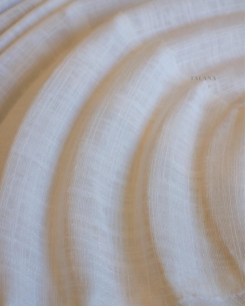 May include: A sheer, off-white fabric featuring a fine, semi-transparent weave with subtle vertical and horizontal lines. The material is draped in soft, flowing curves, creating a layered effect. The word TALANA is printed in small, dark letters.