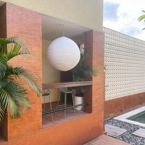 May include: A large, spherical white paper lantern hangs from the ceiling of an outdoor alcove. The alcove features red brick walls, a wooden bar counter, and two metal bar stools. A green palm plant is visible inside the space.