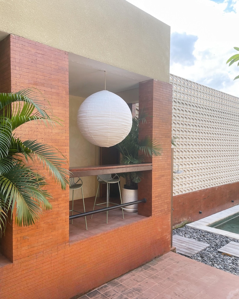 May include: A large, spherical white paper lantern hangs from the ceiling of an outdoor alcove. The alcove features red brick walls, a wooden bar counter, and two metal bar stools. A green palm plant is visible inside the space.