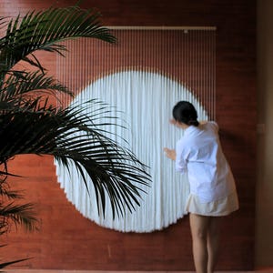 Könnte beinhalten: Ein großer kreisförmiger Wandbehang, der aus zahlreichen vertikalen weißen Baumwollschnüren besteht, die an einer horizontalen Holzstange hängen. Das weiße Faser-Kunstwerk hat eine dichte, geschichtete Textur, die eine Kreisform bildet.