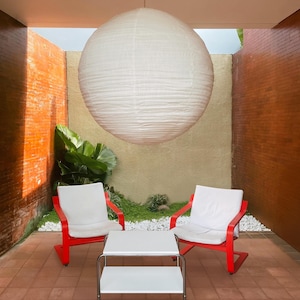 May include: An outdoor patio setting featuring two red chairs with white cushions and a white table with a chrome frame. A large, round, white paper lantern hangs from the ceiling. The patio has brick walls and a tiled floor.