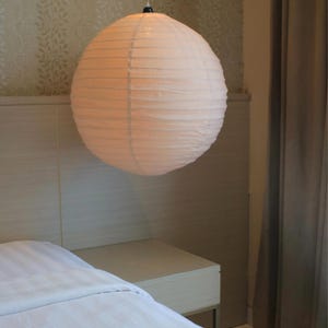 May include: A spherical paper lantern pendant light with a ribbed texture, emitting a soft, warm glow. The white paper shade is suspended from a thin cord, featuring a circular shape that provides diffused illumination in a room setting.