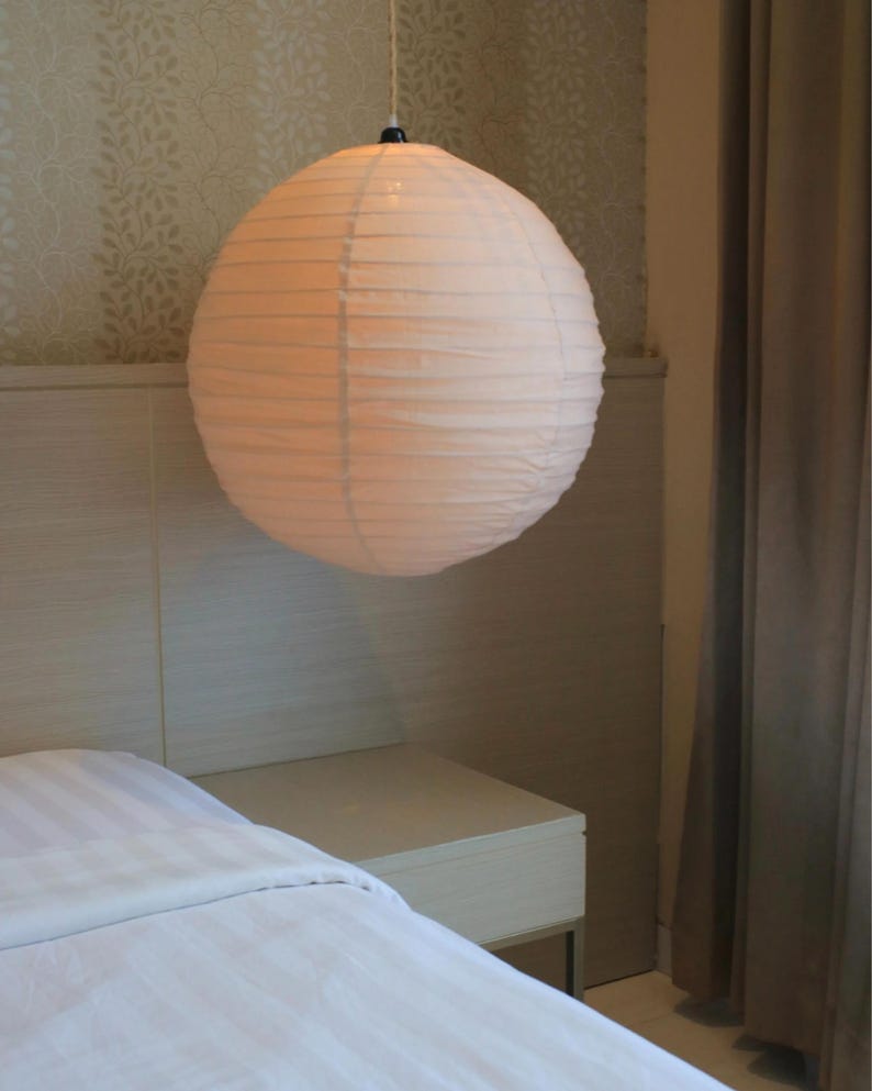 May include: A spherical paper lantern pendant light with a ribbed texture, emitting a soft, warm glow. The white paper shade is suspended from a thin cord, featuring a circular shape that provides diffused illumination in a room setting.
