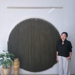 Könnte beinhalten: Ein großer, kreisförmiger Wandbehang mit einem strukturierten, dunkelolivgrünen Design. Das Kunstwerk ist an einer Holzstange aufgehängt und hängt an einer weißen Wand. Eine Frau steht rechts von dem Kunstwerk.