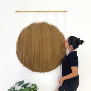 May include: A large, circular wall hanging with a brown, textured design resembling a sun. The artwork is suspended from a wooden rod, with white vertical strands above the brown circle. The background is a plain white wall.