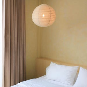 May include: A spherical paper pendant lamp with a ribbed texture hangs from a thin cord. The light fixture emits a soft, warm glow, illuminating the surrounding space with a gentle, diffused light that highlights the delicate paper construction.