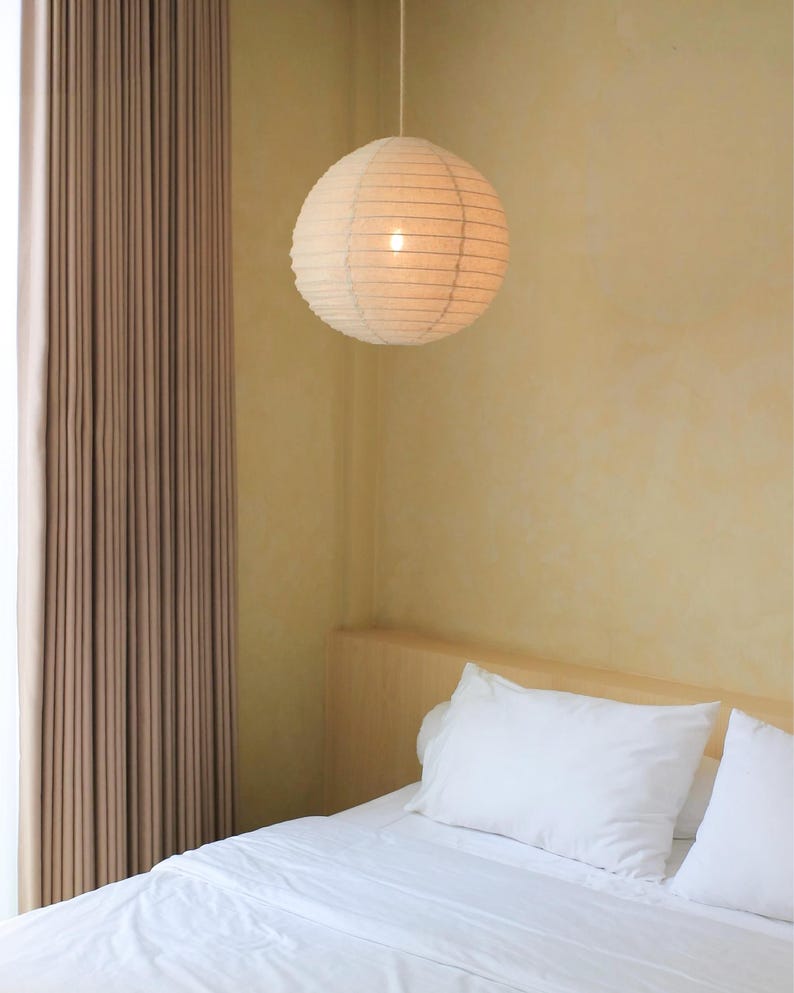 May include: A spherical paper pendant lamp with a ribbed texture hangs from a thin cord. The light fixture emits a soft, warm glow, illuminating the surrounding space with a gentle, diffused light that highlights the delicate paper construction.