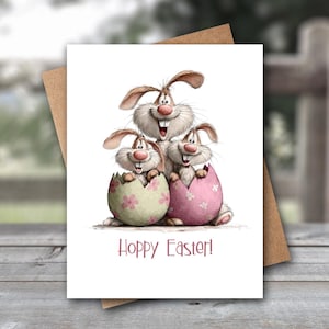 May include: An Easter greeting card featuring three cartoon bunnies. Two bunnies are peeking out of decorated Easter eggs, one pink and one green. The top bunny smiles above the eggs. The words "Hoppy Easter!" are written in red.
