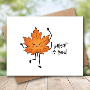 May include: A white greeting card featuring a cartoon orange maple leaf with a smiling face, thin black arms and legs, and the black text "I beleaf in you!" printed to the right of the character. The card is placed against a brown envelope.