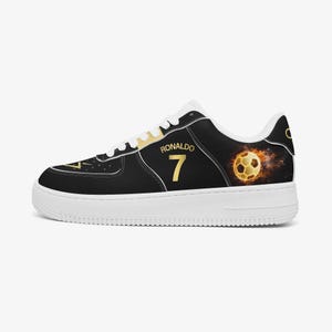 May include: Black and white sneaker with gold accents. The shoe features the name "RONALDO" and the number "7" in gold. A flaming soccer ball graphic is on the side. The shoe has a white sole.