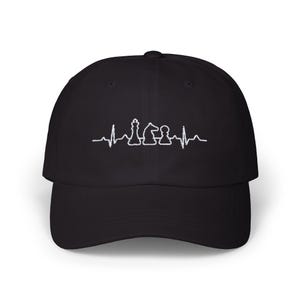 Puede incluir: Gorra de béisbol negra con un diseño bordado en blanco de una línea de latidos del corazón y piezas de ajedrez. Las piezas de ajedrez incluyen un rey, un caballo y un peón. La gorra tiene una visera curva.