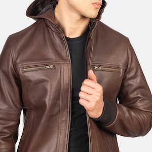 Könnte beinhalten: Eine braune Lederjacke mit Reißverschluss und Kapuze. Die Jacke hat zwei Reißverschlusstaschen auf der Vorderseite und einen gerippten Strickbund am linken Ärmel.