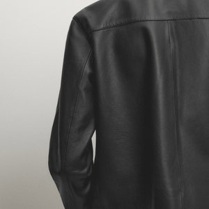May include: A black leather jacket with a collared neckline and long sleeves. The jacket has a simple design and a slightly cropped hem.