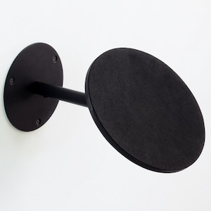 Puede incluir: Un perchero negro para sombreros montado en la pared. El perchero tiene una base circular fijada a la pared con tornillos, un brazo de conexión y una plataforma circular más grande. El diseño es minimalista y moderno.