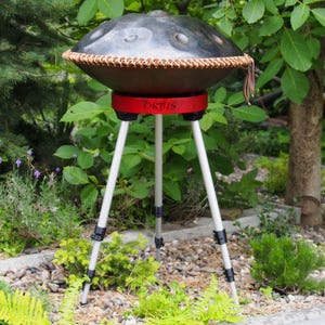Puede incluir: Un handpan de acero con un borde tejido marr&oacute;n y una base roja, sostenido por un tr&iacute;pode blanco. La palabra "ORBIS" est&aacute; impresa en la base roja. El instrumento est&aacute; situado al aire libre, en medio de una exuberante vegetaci&oacute;n y un suelo cubierto de piedras.
